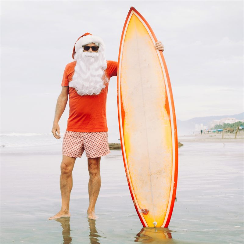 Santa in board shorts holding a surfboard - Christmas in July gift card promo at Escaped In Time